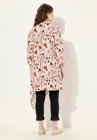 Vloeiende, knielange, bloemen tuniek in crème met rode en paarse bloemen, gedragen over zwarte broeken met kantafwerking. Draag het met lichte espadrilles.