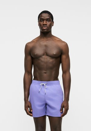 Homme musclé à la peau foncée, debout face à devant, portant un short de bain violet clair avec un cordon blanc sur un fond uni.