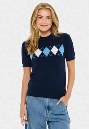 Jeune femme portant un pull à manches courtes bleu marine avec un motif argyle bleu et blanc sur la poitrine, vêtue d'un jean bleu clair, souriant à la caméra.