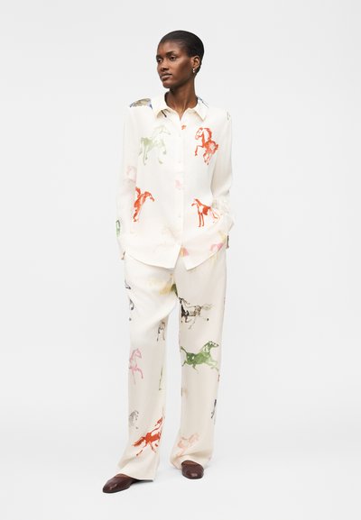 Nainen, jolla on päällään löysä valkoinen paita ja housut, joissa on monivärisiä hevosen kuvia, seisoo kädet taskuissa yksivärisen taustan edessä.