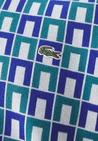 Stickad tröja med ett geometriskt mönster i turkos, blått och vitt, med en liten grön krokodil-logotyp broderad på tyget.