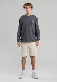 Harmaa pitkähihaisen paita, jossa on pieni grafiikka vasemmassa rinnassa, yhdistettynä kermaisiin lyhyisiin shortseihin. Mallilla on päällä valkoiset sneakerit.