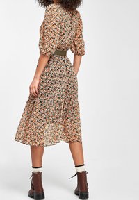Mujer de pie, con la espalda visible, vistiendo un vestido midi floral en naranja y negro, calcetines blancos y botas marrones con cordones, sobre un fondo liso.
