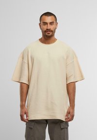 Tricou oversized bej, cu mânecă scurtă, realizat dintr-un material texturat, având un guler rotund și detalii brodate subtile pe față.