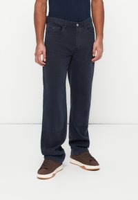 Muž stojaci na jednofarebnom bielom pozadí, oblečený v tmavomodrých cargo nohaviciach so bočnými vreckami a hnedých teniskách.