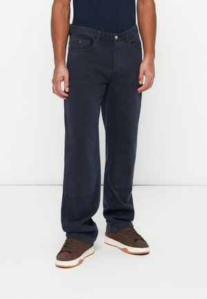 Mann in dunkelblauen Cargo-Hosen mit seitlichen Taschen und braunen Sneakers, der vor einem schlichten weißen Hintergrund steht.