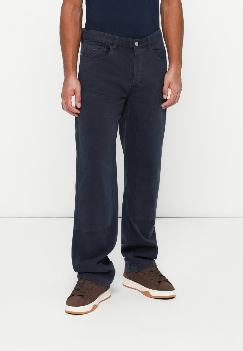 Muž stojaci na jednofarebnom bielom pozadí, oblečený v tmavomodrých cargo nohaviciach so bočnými vreckami a hnedých teniskách.