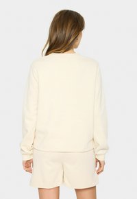 Sudadera color crema con un corte relajado, mangas largas y puños acanalados, combinada con pantalones cortos a juego. Textura suave, diseño minimalista.