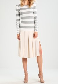 Top a maniche lunghe a righe grigie e bianche con costine abbinato a una gonna plissettata beige con aperture laterali. Il modello indossa tacchi a cinturino nudo.