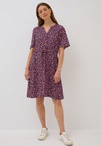 Kurzärmliges Kleid in dunkelviolett mit weißem Blättermuster, V-Ausschnitt, verstellbarem Bund mit Kordelzug, knielang, kombiniert mit weißen Sneakern.