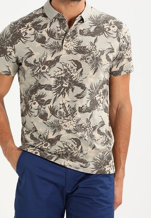 Homme portant un polo à manches courtes beige à imprimé floral foncé et un pantalon bleu, debout avec une main dans la poche.