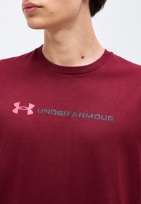 Νεαρό άτομο φοράει μπορντό μπλούζα Under Armour με ροζ λογότυπο και κείμενο, ορατά από το λαιμό μέχρι το άνω μέρος του στήθους.