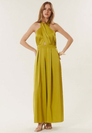 Femme portant une combinaison sans manches couleur olive à col halter, jambes larges, et des sandales à talons hauts beige, debout les mains sur les hanches.