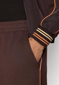 Καφέ αθλητικό παντελόνι jogging με ελαστική μέση και πλαϊνή τσέπη. Η μανσέτα έχει ριγέ διακοσμήσεις σε πορτοκαλί, εκρού και σκούρο καφέ. Λείο υφή.