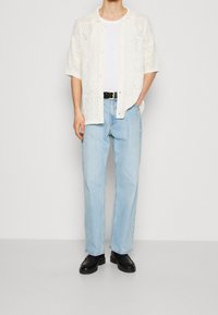 Lichte, open-gehaakte crèmekleurige cardigan over een wit T-shirt, gecombineerd met lichtblauwe rechtlijnige jeans en zwarte schoenen.