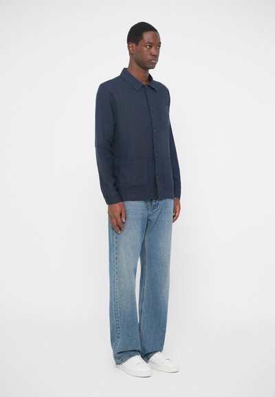 Moški stoji, oblečen v temno modro srajco s gumbi, svetlo modre kavbojke in bele superge proti enostavni beli podlagi.