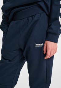 Blå joggerbyxor av mjukt tyg med elastisk midjeresår, sidofickor och en vit logga på underbenet.