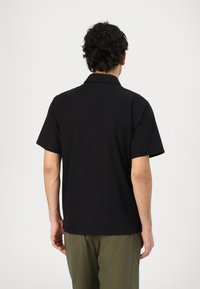 Čierna košeľa s krátkym rukávom a golierom, vyrobená z hladkej látky, priliehavý strih a rovný lem, nosené s olivovozelenými nohavicami.