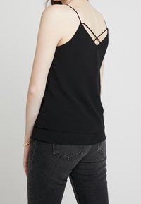Camisola nera con spalline sottili e dettaglio incrociato sulla schiena. Tessuto liscio, vestibilità comoda, orlo a strati, abbinata a jeans grigio scuro.