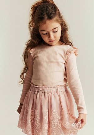 Junges Mädchen mit langem lockigem Haar, trägt ein pinkfarbenes Langarmshirt und einen pinken Rock mit Spitzenbesatz, schaut nach unten und hält den Saum ihres Rocks.