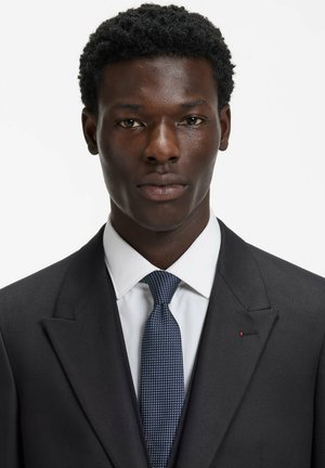 Black suit jacket with a textured white shirt, and a blue patterned tie. Notable detailing includes a red accent on the lapel.