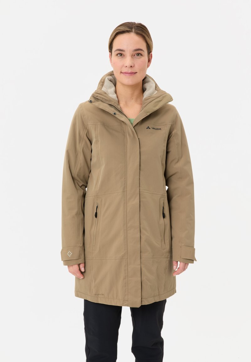 Donna che indossa un giubbotto con cappuccio color sabbia a lunghezza ginocchio con tasche con zip e pantaloni neri, in piedi su uno sfondo chiaro e uniforme.