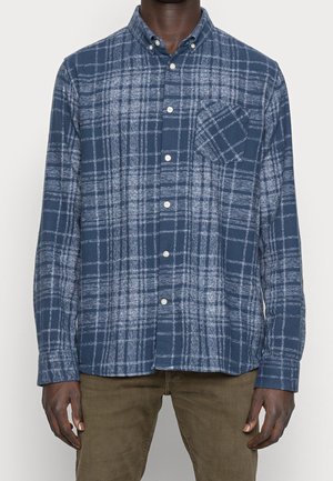 Muž v modro-bílé kárované košili s dlouhým rukávem a náprsní kapsou a v hnědých kalhotách stojí před jednobarevným pozadím.