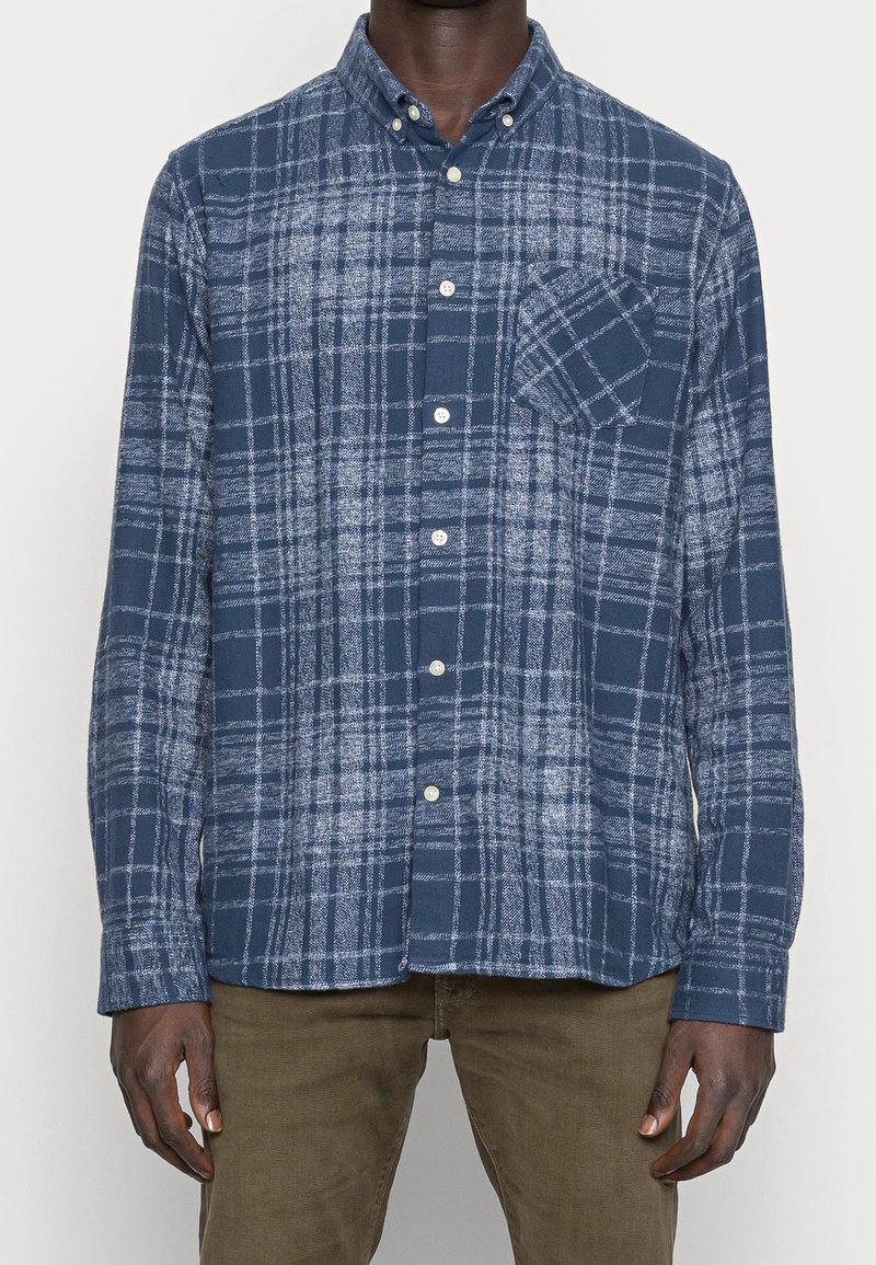 Homme portant une chemise à manches longues à carreaux bleus et blancs avec une poche poitrine et un pantalon marron, debout devant un fond uni.