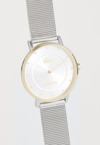 Sudraba sieta josta pulkstenis ar baltu ciparu, zelta toņa rāmis un smalkiem punktu stundu rādītājiem. Lacoste logo izvietots zem 12. stundas pozīcijas.