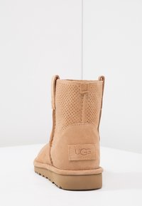 Beżowe zamszowe botki UGG z perforowaną cholewką, masywną podeszwą oraz przeszywaną metką marki na pięcie, wyeksponowane na białej powierzchni i tle.