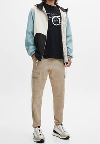 Person, der beige Cargo-Hosen, weiße und schwarze Sneaker, ein schwarzes Grafik-T-Shirt und eine farblich blockierte Jacke mit hellblauen Ärmeln und beigem Körper trägt.