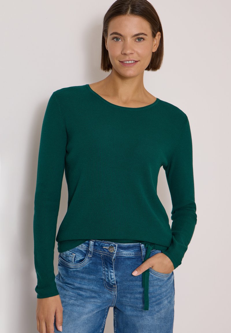 Jeune femme aux cheveux courts et bruns portant un pull vert foncé à manches longues et un jean bleu, une main dans la poche, sur un fond uni.
