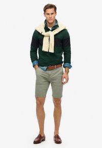 Pullover a righe verdi e blu navy, camicia di denim azzurro chiaro, pantaloni corti color oliva, cintura marrone e mocassini. Maglione in maglia appoggiato sulle spalle.