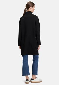 Schwarzer, übergroßer Mantel mit hohem Kragen und seitlichen Taschen, kombiniert mit blauen Jeans und weißen Turnschuhen, von hinten präsentiert.