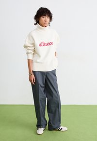 Hvit strikket turtleneck genser med rødt 'ellesse' logo, sammen med løse grå bukser. Hvite joggesko med svarte striper. Grønn gulv bakgrunn.