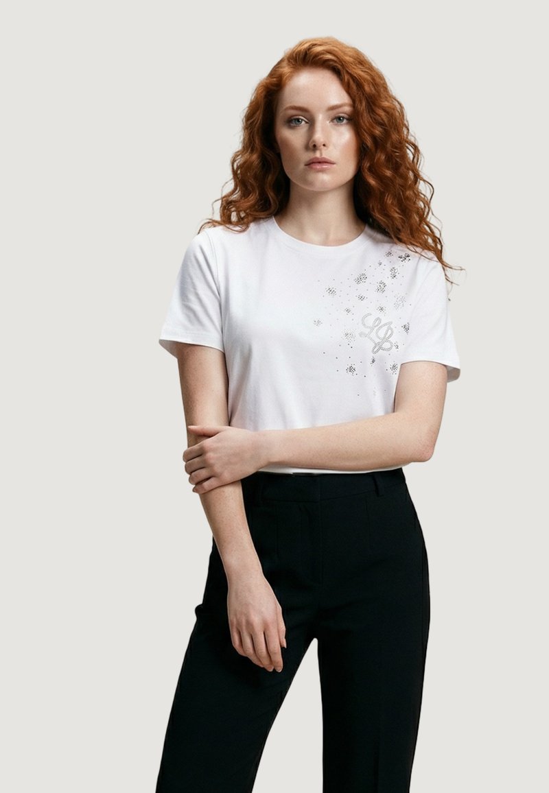 Joven con cabello largo y rizado de color rojo, vestida con una camiseta blanca con un diseño sutil en el pecho y pantalones negros, de pie frente a un fondo liso.