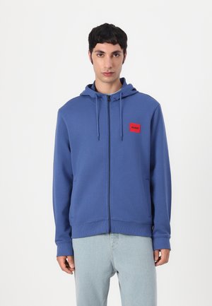 Hoodie bleu zippé avec une petite patch logo rouge sur la poitrine. Comprend des cordons, des poches avant et une texture de tissu douce.
