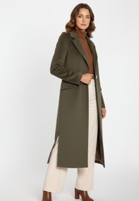 Olijfgroene lange jas met een gestructureerd ontwerp, zijopeningen en een voering met luipaardprint. Draagt over een bruine coltrui en beige broek.