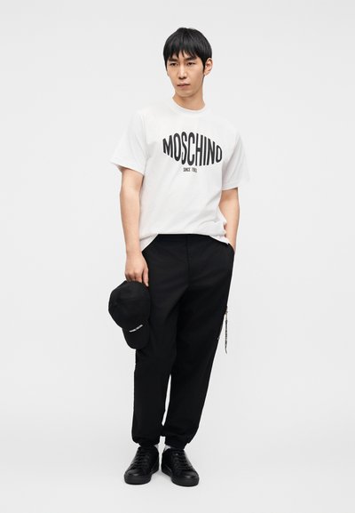 Λευκό κοντομάνικο μπλουζάκι με μαύρο κείμενο "MOSCHINO", σε συνδυασμό με μαύρο παντελόνι και μαύρο καπέλο που κρατιέται στο χέρι. Μαύρα αθλητικά παπούτσια ολοκληρώνουν την εμφάνιση.