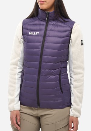 Femme portant un gilet matelassé violet sans manches avec le logo « MILLET » sur un haut à manches longues gris clair, pantalon beige, debout devant un fond blanc.
