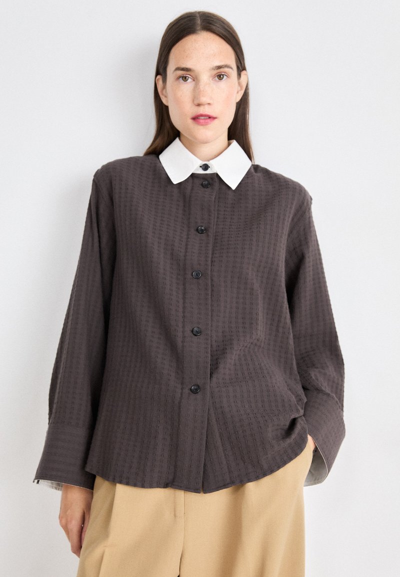 Dark brown textured button-up shirt with a white collar, featuring black buttons and long sleeves, styled with relaxed fit and subtle pattern.