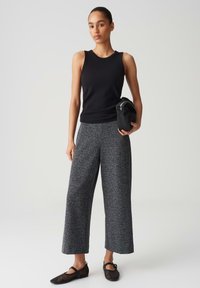 Schwarzes ärmelloses Top kombiniert mit locker sitzenden, grauen gemusterten Hosen. Das Model hält eine schwarze Tasche. Einfaches, modernes Design mit einer entspannten Passform.