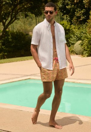 Muž nosící sluneční brýle, bílou krátkosrstou košili a vzorované plavky kráčí bos po boku bazénu v slunečný den.