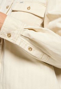 Camicia button-up color crema realizzata in tessuto a spina di pesce. Presenta bottoni arrotondati e due tasche sul petto con etichetta.