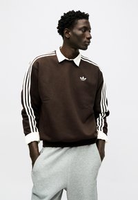 Νεαρός άνδρας με καφέ φούτερ Adidas με λευκές ρίγες και γκρι φόρμα, στέκεται με τα χέρια στις τσέπες, κοιτάζοντας προς τα πλάγια.
