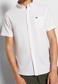 Vitt kortärmat skjorta med krage, med en bröstficka på vänster sida och en liten marinblå krona-logotyp. Slät textur.