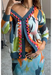 Sweterek na drutach ma kolorowy wzór kwiatowy z akcentami w postaci pomponów z przodu. Niebieska baza z odcieniami różu, pomarańczy i zieleni.