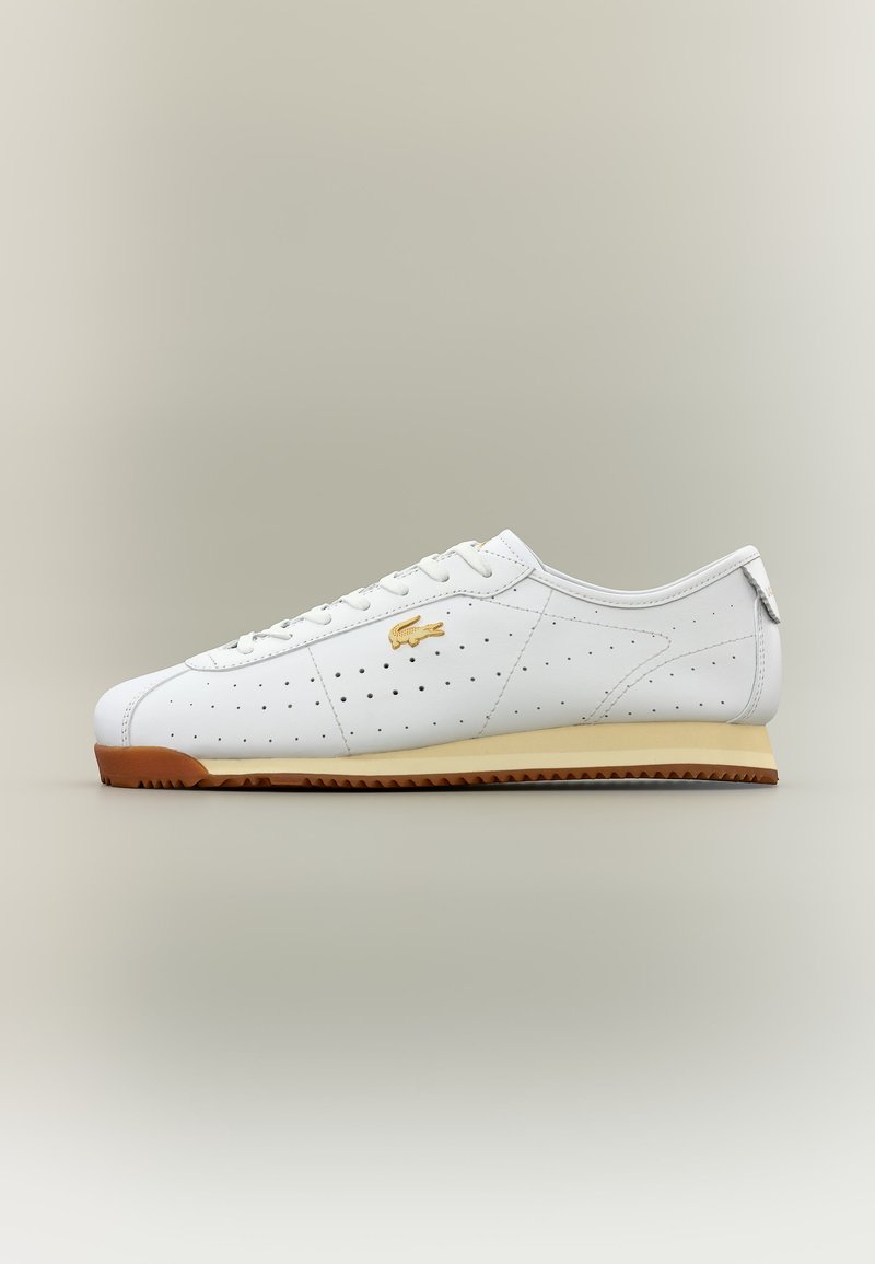 Λευκό sneaker χαμηλού προφίλ με διάτρητο δέρμα, χρυσλό λογότυπο στη πλευρά, μπεζ ενδιάμεση σόλα και καφέ ραβδωτή εξωτερική σόλα σε ουδέτερο φόντο.