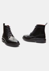 Botas de cuero negro con cordones y un diseño tipo brogue, que presentan detalles perforados, una lengüeta en la parte trasera y una suela texturizada para mayor agarre.