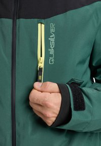 Grüne und schwarze Jacke mit einer Reißverschlusstasche, die einen gelben Zippergriff hat. Textlogo auf der Vorderseite, mit Klettverschlüssen an den Manschetten. Glatte Stoffstruktur.